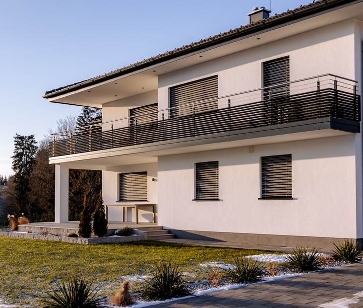 Modernes zweistöckiges Haus mit weißer Fassade, Balkon mit Metallgeländer und Fenstern mit Rollläden, davor Rasen und Weg.