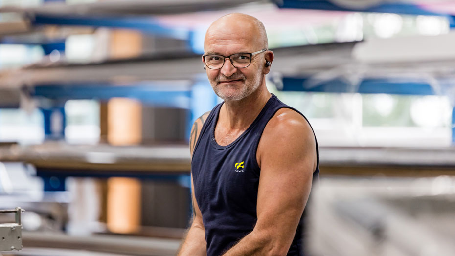 Ein Mann mit Glatze und Brille trägt eine ärmellose Arbeitskleidung und steht vor einer industriellen Produktionsumgebung. Er lächelt in die Kamera, während er eine Maschine bedient. Der Hintergrund zeigt Regale und Produktionsanlagen. Mann in ärmelloser Arbeitskleidung in einer Produktionshalle