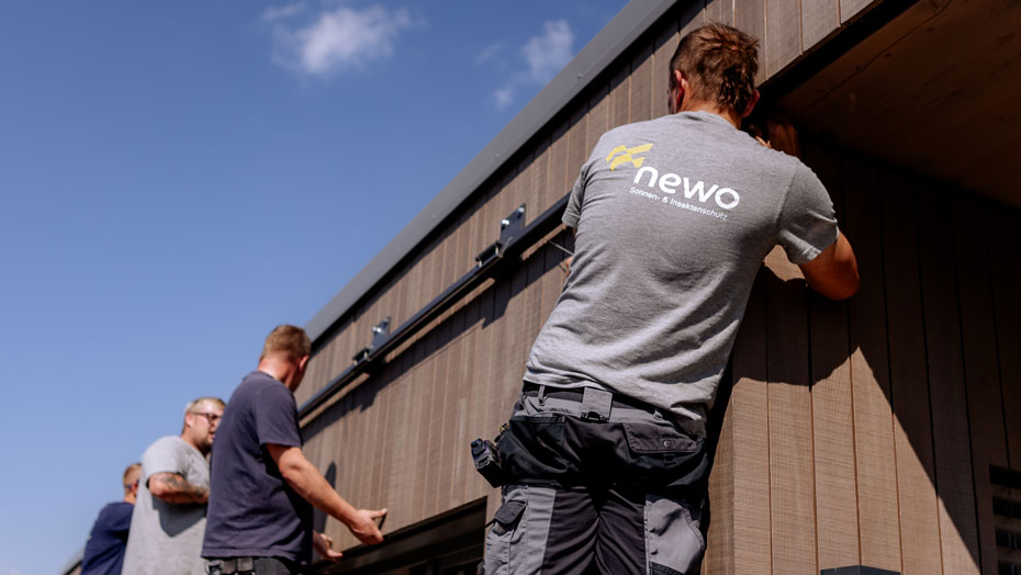 Mehrere Arbeiter installieren eine Markise im Freien. Die Person im Vordergrund trägt ein graues T-Shirt mit dem Logo 'newo'. Arbeiter installieren eine Markise im Freien, eine Person trägt ein T-Shirt mit einem Logo.