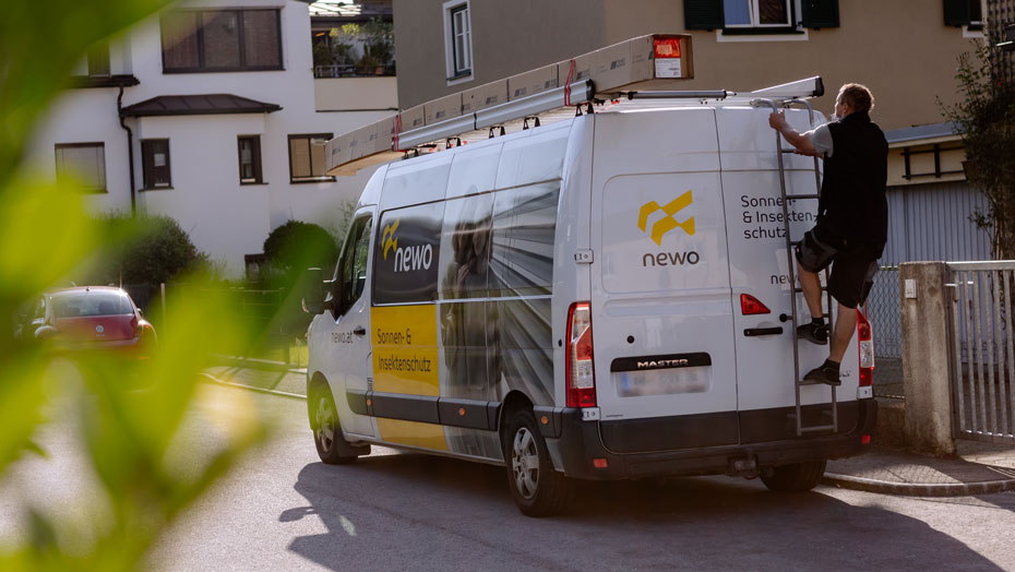 Ein Mann in Arbeitskleidung klettert auf eine Leiter am Heck eines weißen Transporters mit der Aufschrift 'newo' und 'Sonnen- & Insektenschutz'. Der Transporter steht in einer Wohngegend. Mann klettert auf Leiter am Transporter mit 'newo' Beschriftung in Wohngebiet