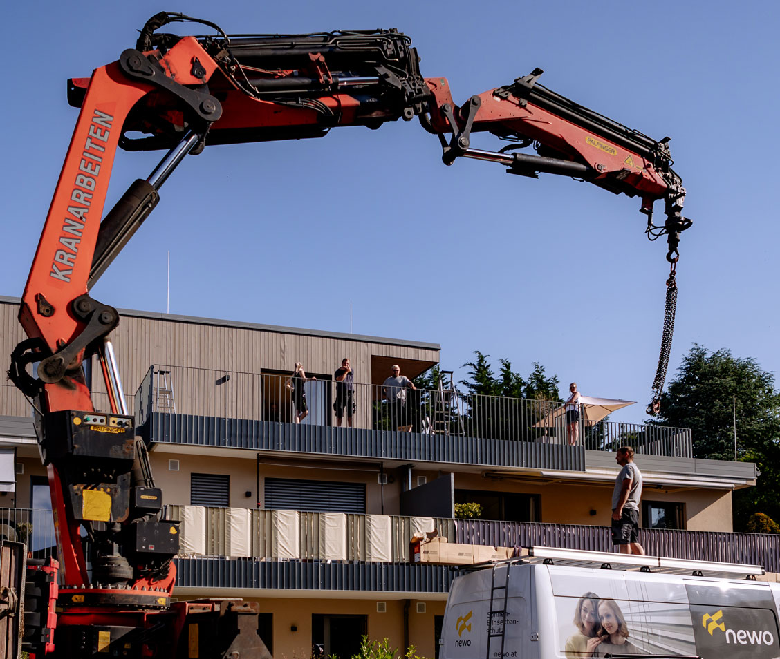 Kran hebt Last vor einem Wohngebäude, Personen beobachten vom Balkon, Mann auf Lieferwagendach.