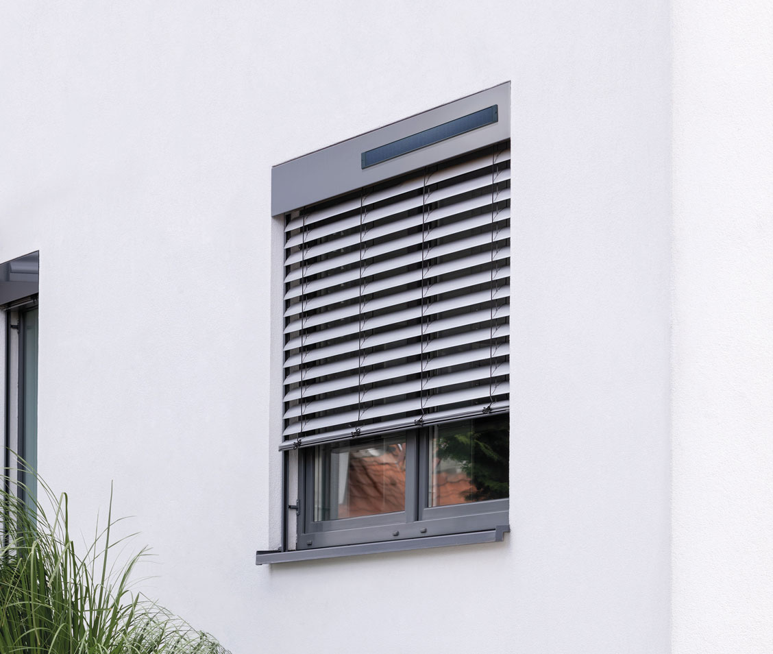 Fenster mit grauem Rahmen und Solar-Raffstore an weißer Hauswand