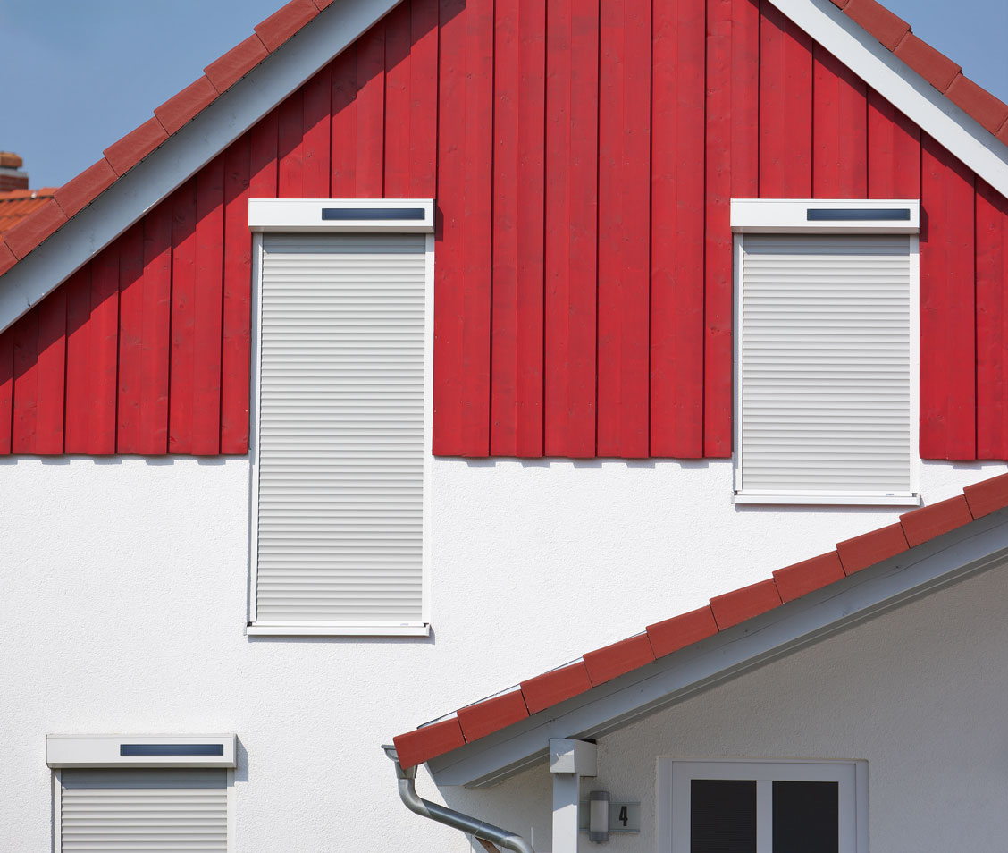 Hausfassade mit roter Holzverkleidung und geschlossenen weißen Solar-Rollläden an den Fenstern.